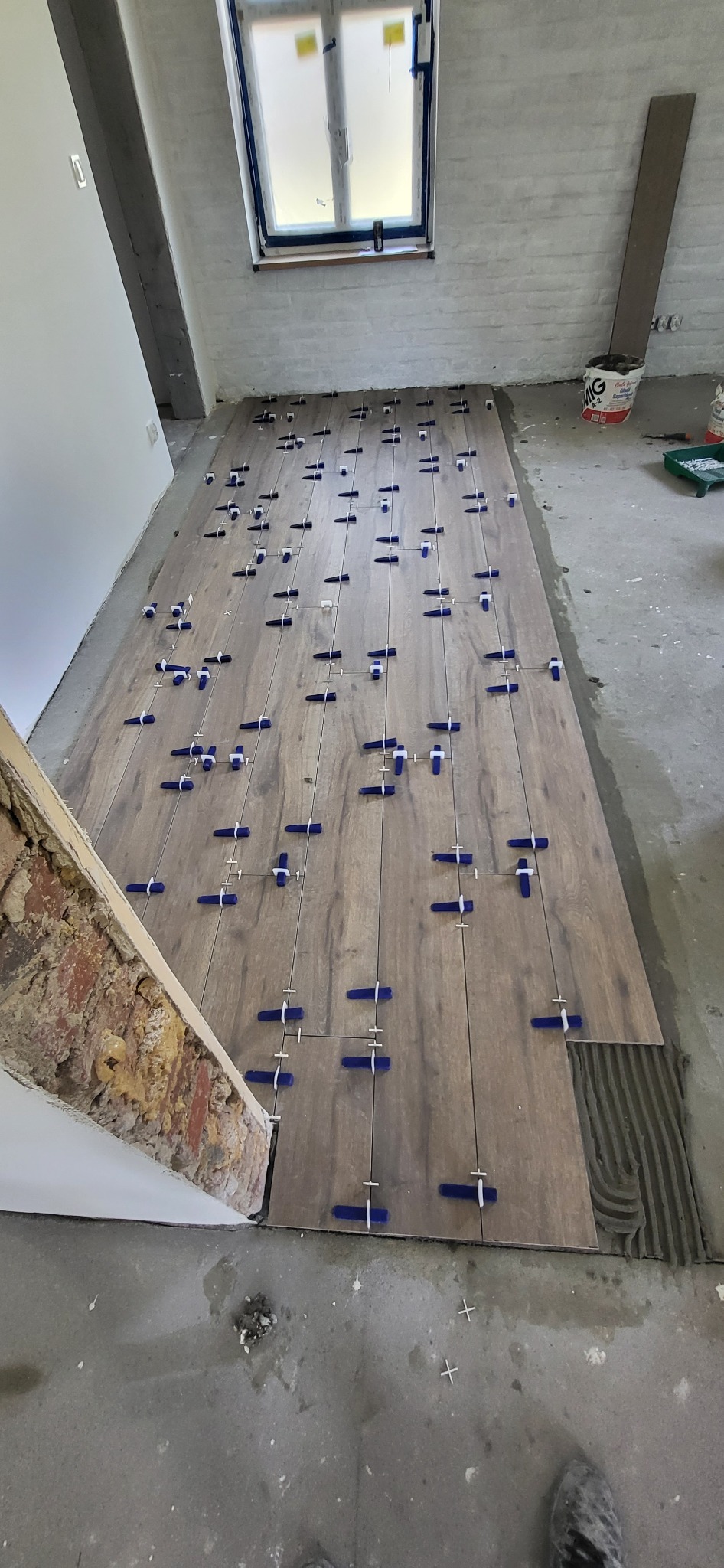 Układanie płytek podłogowych imitujących drewno z użyciem niebieskich krzyżyków dystansowych; widoczny fragment ściany z cegły i resztki zaprawy na podłodze.