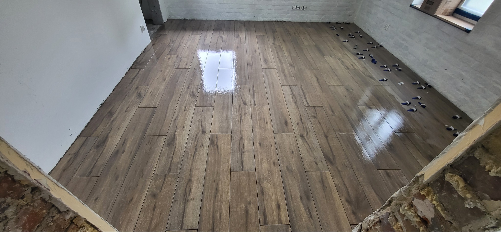 Świeżo ułożona, błyszcząca podłoga z płytek imitujących drewno, z widocznymi niebieskimi klinami poziomującymi na krawędziach, w pomieszczeniu z surowymi ścianami z cegły i betonu.
