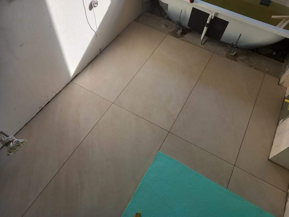 Ułożone beżowe płytki podłogowe w łazience w trakcie remontu, widoczna wanna, nieukończone ściany i fragment niebieskiej płyty izolacyjnej.