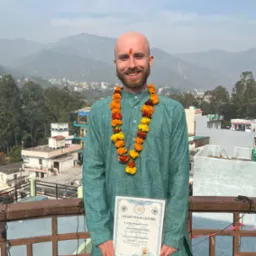 Uśmiechnięty mężczyzna z tilaką na czole, w zielonej szacie i girlandzie z pomarańczowych kwiatów, trzymający certyfikat Anadi Yoga Centre na tle górskiego krajobrazu i zabudowań miejskich.