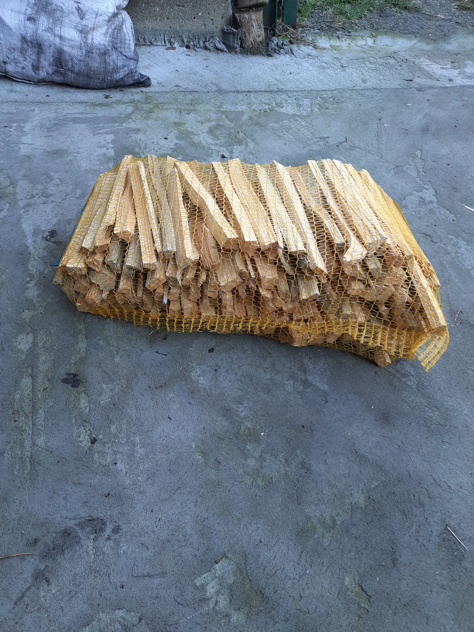 Worek z rąbanym drewnem kominkowym w siatce, leżący na szarym betonowym podłożu, z widocznym workiem z węglem w tle.