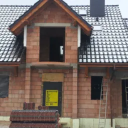 Budynek w trakcie budowy z częściowo pokrytym dachem dachówką ceramiczną, widoczne rusztowania, drabina i palety z dachówką przed budynkiem.