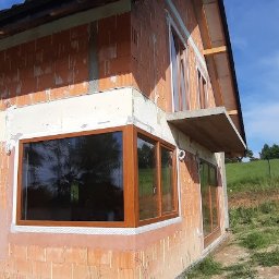 INVEST-DOM - Montaż okien drewnianych w surowym budynku z czerwonej cegły, widoczne detale uszczelnienia i pianki montażowej, zarys sylwetki fotografa w dolnej części kadru.