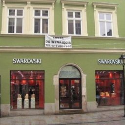 INVEST-DOM - Fasada budynku w pistacjowym kolorze z trzema białymi oknami na górze i dwoma dużymi witrynami sklepowymi na dole, nad którymi widnieje napis 'SWAROVSKI', a nad nimi baner 'Lokal do wynajęcia'.