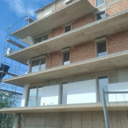 INVEST-DOM - Wielopiętrowy budynek w trakcie budowy, widoczne surowe ściany z cegły, zamontowane okna z roletami zewnętrznymi oraz częściowo ocieplone ściany, rusztowania i materiały budowlane w otoczeniu.