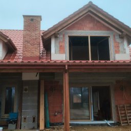 INVEST-DOM - Budowa domu jednorodzinnego w trakcie montażu stolarki okiennej i drzwiowej, widoczne surowe mury, niezabudowane otwory okienne i drzwiowe, ceglany komin, drewniana konstrukcja zadaszenia tarasu.