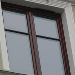 INVEST-DOM - Dwuskrzydłowe okno z brązowymi ramami, biała ozdobna rama okienna, widok z dołu, szare niebo odbija się w szybach.