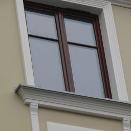 INVEST-DOM - Dwuszybowe okno drewniane z brązowymi ramami i białym ozdobnym obramowaniem na beżowej ścianie budynku, widok z dołu.