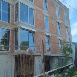 INVEST-DOM - Budynek w trakcie budowy z zamontowanymi oknami i roletami zewnętrznymi, widoczne elementy konstrukcyjne i rusztowania.