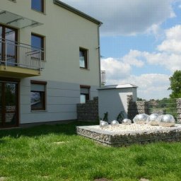 INVEST-DOM - Elewacja budynku z oknami z roletami zewnętrznymi, balkonem z szklanym zadaszeniem oraz dekoracyjnym gabionem z metalowymi kulami na trawniku.