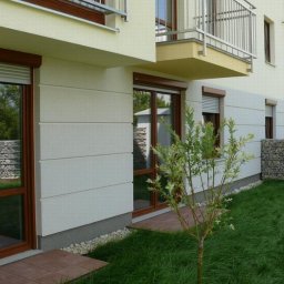 INVEST-DOM - Elewacja budynku z oknami z brązowymi ramami i częściowo opuszczonymi roletami zewnętrznymi, widok z niskiej perspektywy na trawnik i małe drzewko.