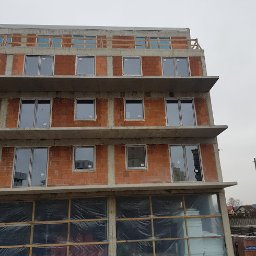 INVEST-DOM - Surowy budynek wielorodzinny w trakcie montażu okien PCV i aluminiowych, widoczne okna z szarymi ramami na tle ceglastego muru, dolne okna zabezpieczone folią, w tle pochmurne niebo.