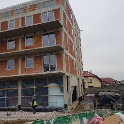 INVEST-DOM - Budynek w trakcie budowy z zamontowanymi oknami, widoczny pracownik w kamizelce odblaskowej i kontener na śmieci.