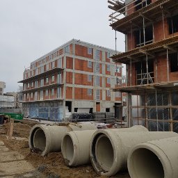 INVEST-DOM - Budynek w trakcie budowy z widocznymi oknami PCV i betonowymi rurami na pierwszym planie, szare niebo w tle.