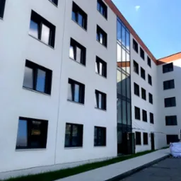 Elewacja budynku wielopiętrowego z regularnie rozmieszczonymi oknami z czarnymi ramami, widoczne odbicia w szybach, chodnik i trawnik przed budynkiem.