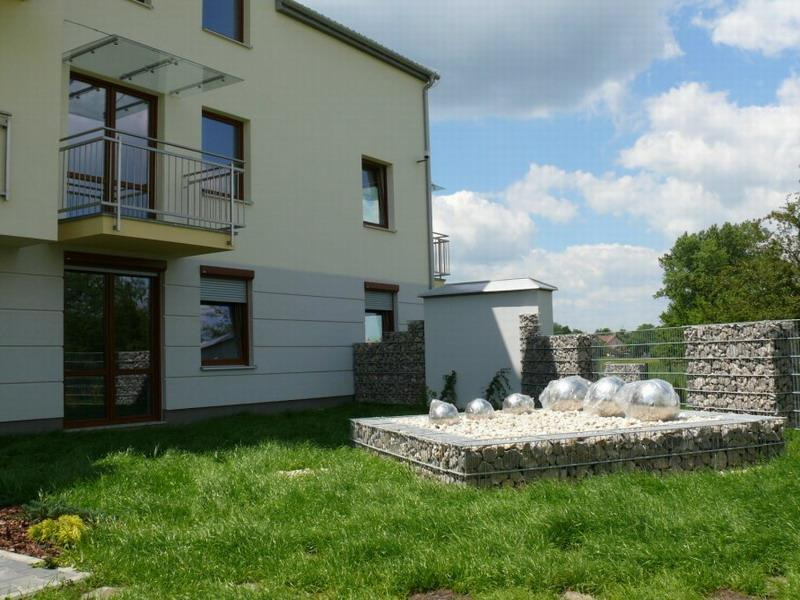 Elewacja budynku z oknami z roletami zewnętrznymi, balkonem z szklanym zadaszeniem oraz dekoracyjnym gabionem z metalowymi kulami na trawniku.