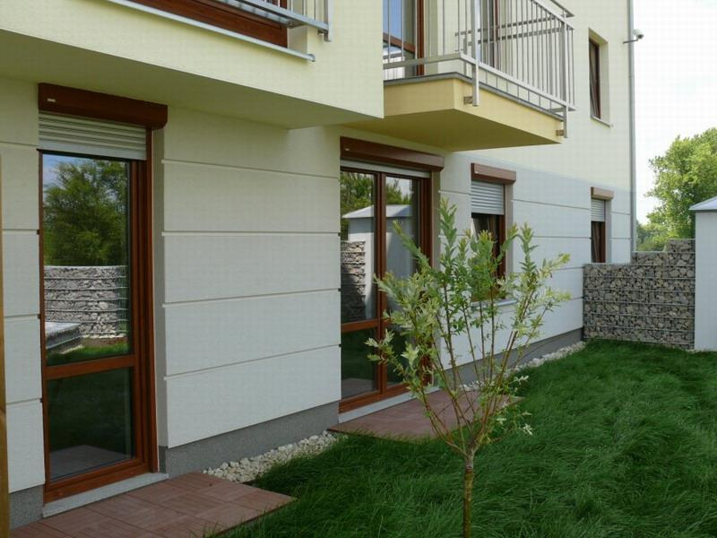 Elewacja budynku z oknami z brązowymi ramami i częściowo opuszczonymi roletami zewnętrznymi, widok z niskiej perspektywy na trawnik i małe drzewko.