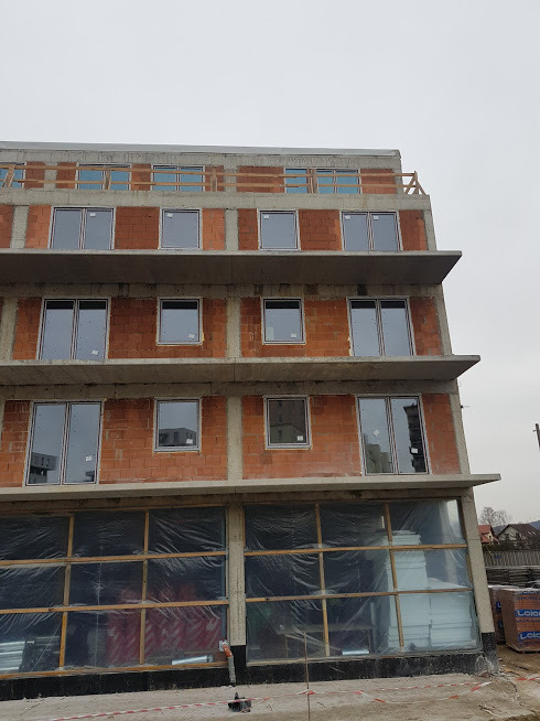 Surowy budynek wielorodzinny w trakcie montażu okien PCV i aluminiowych, widoczne okna z szarymi ramami na tle ceglastego muru, dolne okna zabezpieczone folią, w tle pochmurne niebo.