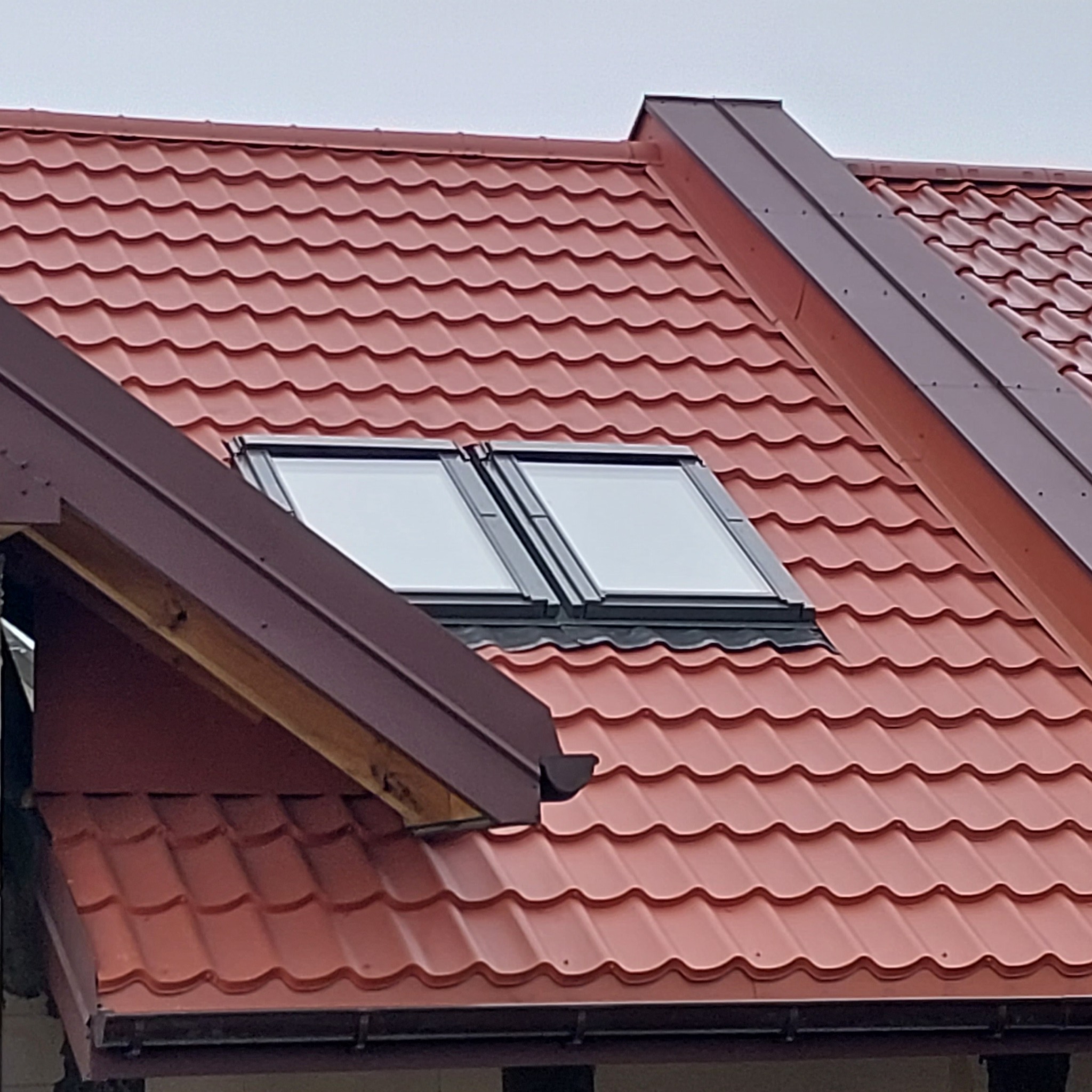 Czerwony dach z blachy trapezowej z dwoma oknami dachowymi i brązowymi obróbkami blacharskimi.