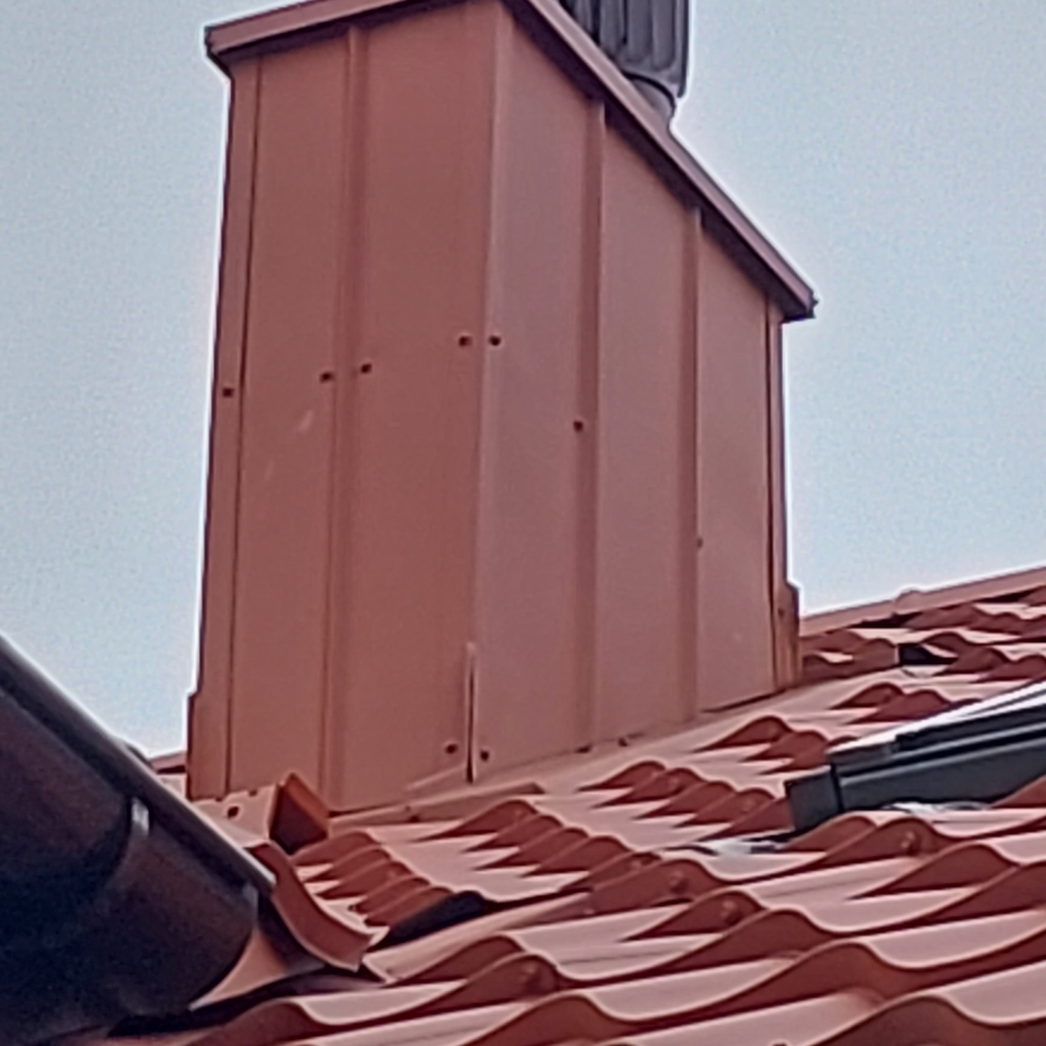 Nowy, brązowy komin obłożony blachą na dachu z dachówki ceramicznej w kształcie fali.