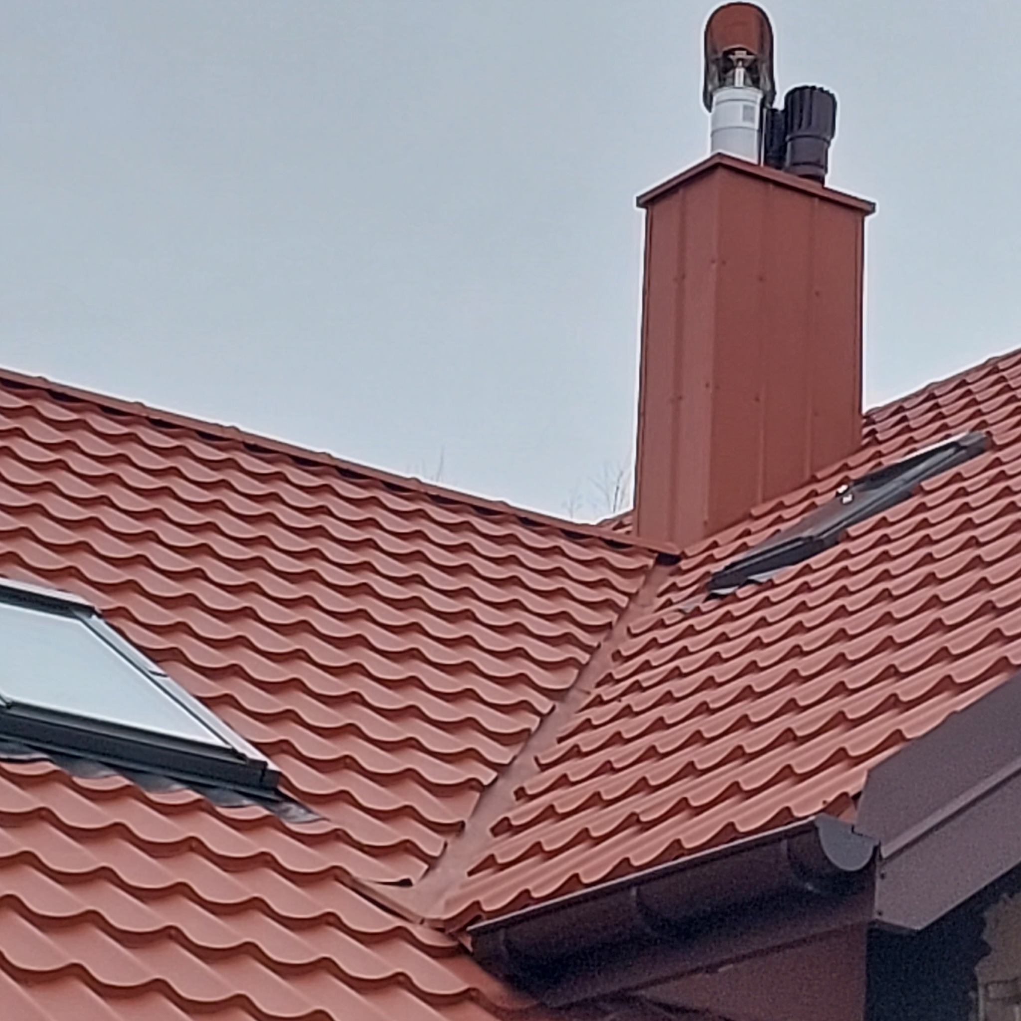 Czerwony dach z blachodachówki z oknem dachowym i kominem z nasadami wentylacyjnymi