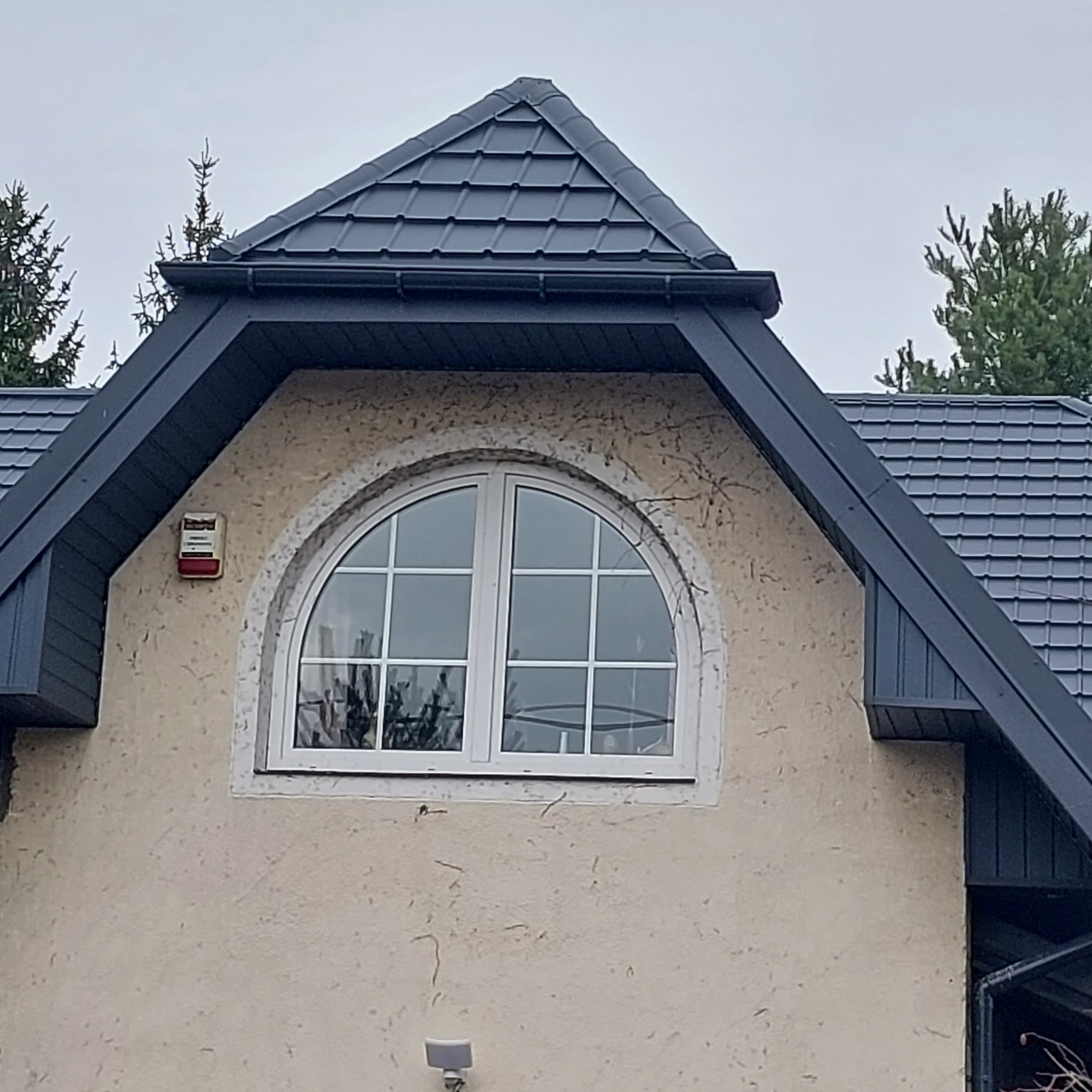 Dom z ciemną dachówką i obróbkami blacharskimi, widoczny fragment elewacji z oknem w kształcie półkola i alarmem.