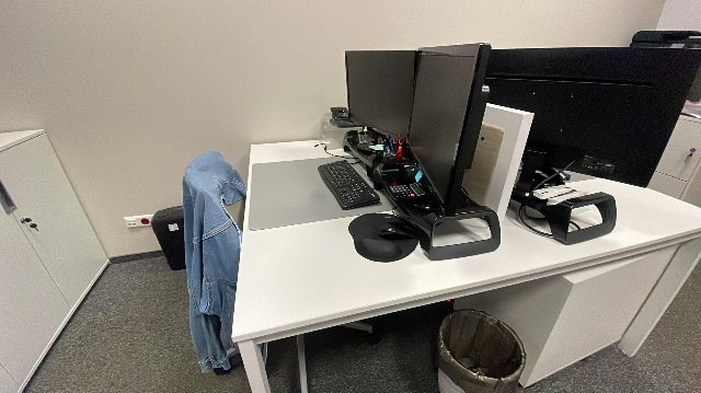 Białe biurko z dwoma monitorami, klawiaturą i myszką komputerową w biurze. Pod biurkiem widoczny kosz na śmieci. Na krześle wisi jeansowa kurtka.