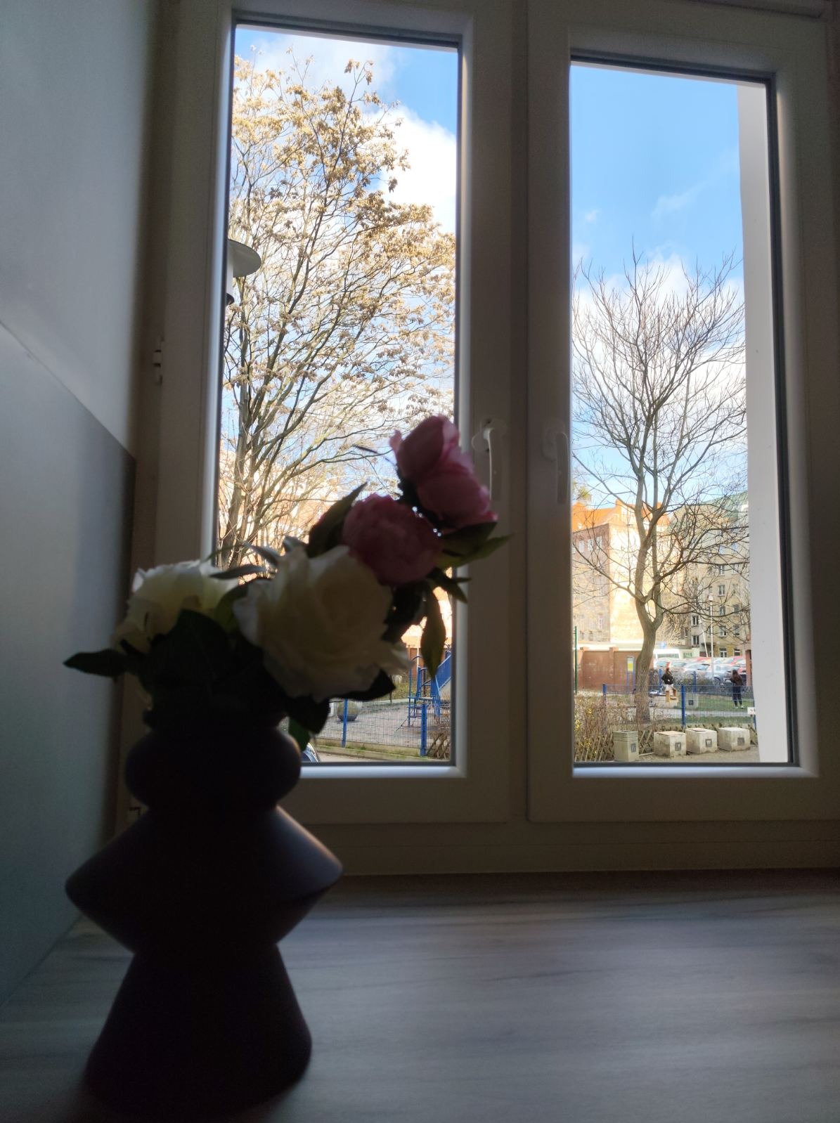 Widok z okna na plac zabaw i drzewa, z wazonem z kwiatami w pierwszym planie. Okno umyte, brak smug. Lekkie odbicie światła na szybie.