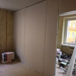 Wnętrze pokoju w trakcie prac wykończeniowych: ściany pokryte płytami gipsowo-kartonowymi, widoczne worki z gipsem, okno z widokiem na budynek, stare ściany z resztkami tapet.