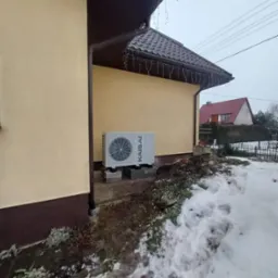 Zewnętrzna jednostka klimatyzacji Kaisai zamontowana na betonowych bloczkach przy elewacji domu, z widocznymi śladami topniejącego śniegu.