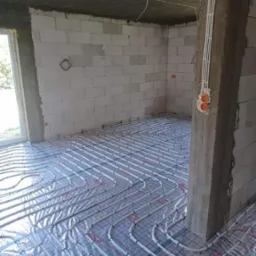 Montaż rur ogrzewania podłogowego na folii z siatką w surowym pomieszczeniu z betonowym słupem i przewodami elektrycznymi.