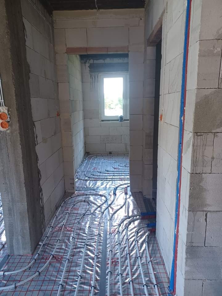 Wnętrze w trakcie instalacji ogrzewania podłogowego, widoczne rury grzewcze na folii z siatką, ściany z betonu komórkowego, okno w głębi pomieszczenia.