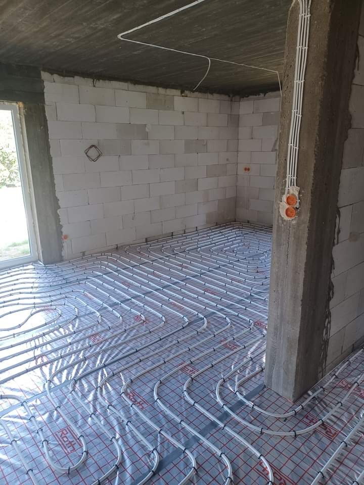 Montaż rur ogrzewania podłogowego na folii z siatką w surowym pomieszczeniu z betonowym słupem i przewodami elektrycznymi.