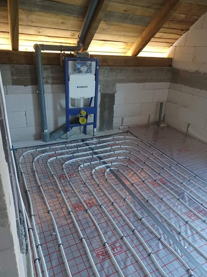 Instalacja ogrzewania podłogowego w surowym pomieszczeniu poddasza, widoczne rury Roth ułożone na folii z kratką, stelaż podtynkowy Geberit oraz nieotynkowane ściany z bloczków.