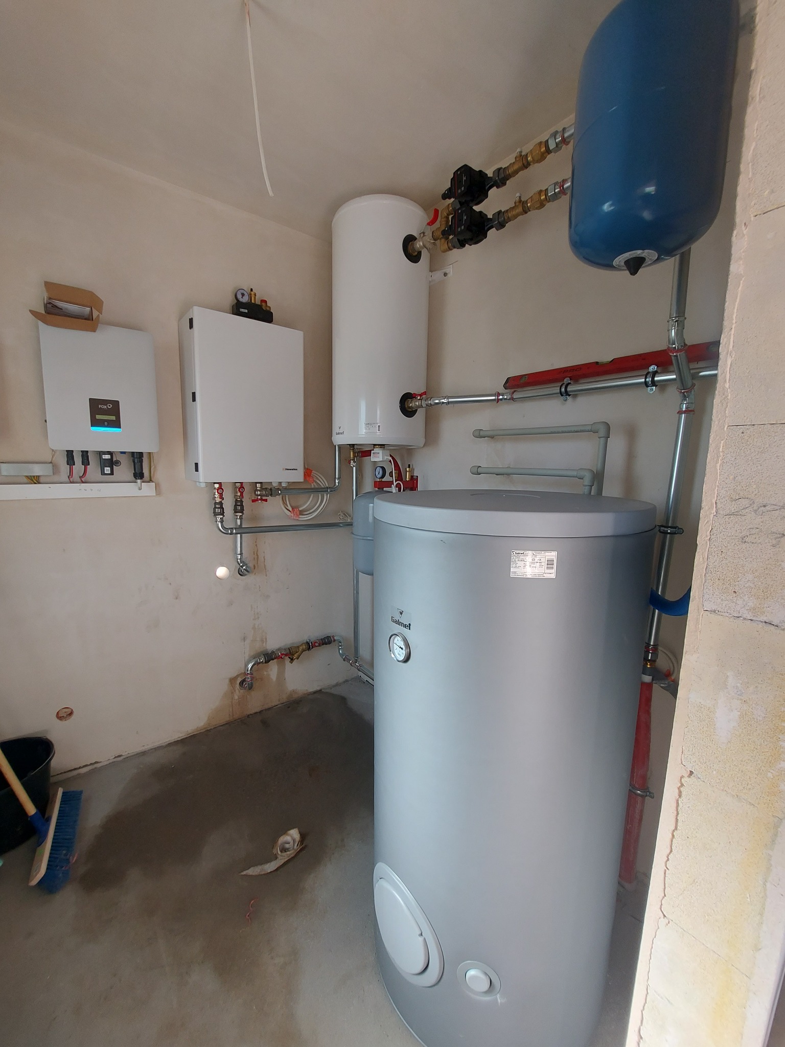 Kotłownia w trakcie instalacji: szary zbiornik Galmet, biały zasobnik, niebieskie naczynie przeponowe, dwa białe panele sterujące z podłączeniami, liczne rury miedziane i stalowe, poziomica...