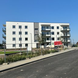 L.T.consulting - Nowoczesny, trzypiętrowy budynek mieszkalny z balkonami, wykończony na biało z szarymi akcentami, widoczny z poziomu nowej asfaltowej drogi i chodnika, z ogrodzeniem tymczasowym i roślinnością...