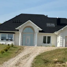 Elegancki dom jednorodzinny z białą elewacją, czarnym dachem z dachówki i podjazdem z jasnego żwiru, otoczony zielonym trawnikiem i niskimi krzewami.