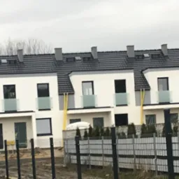Szereg nowo wybudowanych, ocieplonych domów w zabudowie szeregowej z szarą dachówką i elewacją w kolorze złamanej bieli, widoczne żółte rury przy elewacji i ogrodzenie.