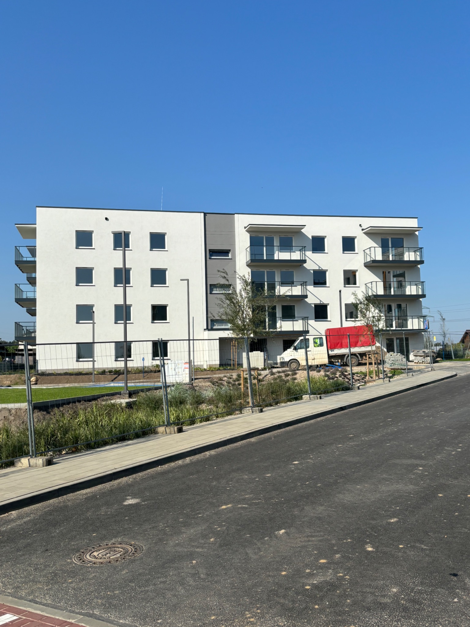 Nowoczesny, trzypiętrowy budynek mieszkalny z balkonami, wykończony na biało z szarymi akcentami, widoczny z poziomu nowej asfaltowej drogi i chodnika, z ogrodzeniem tymczasowym i roślinnością...