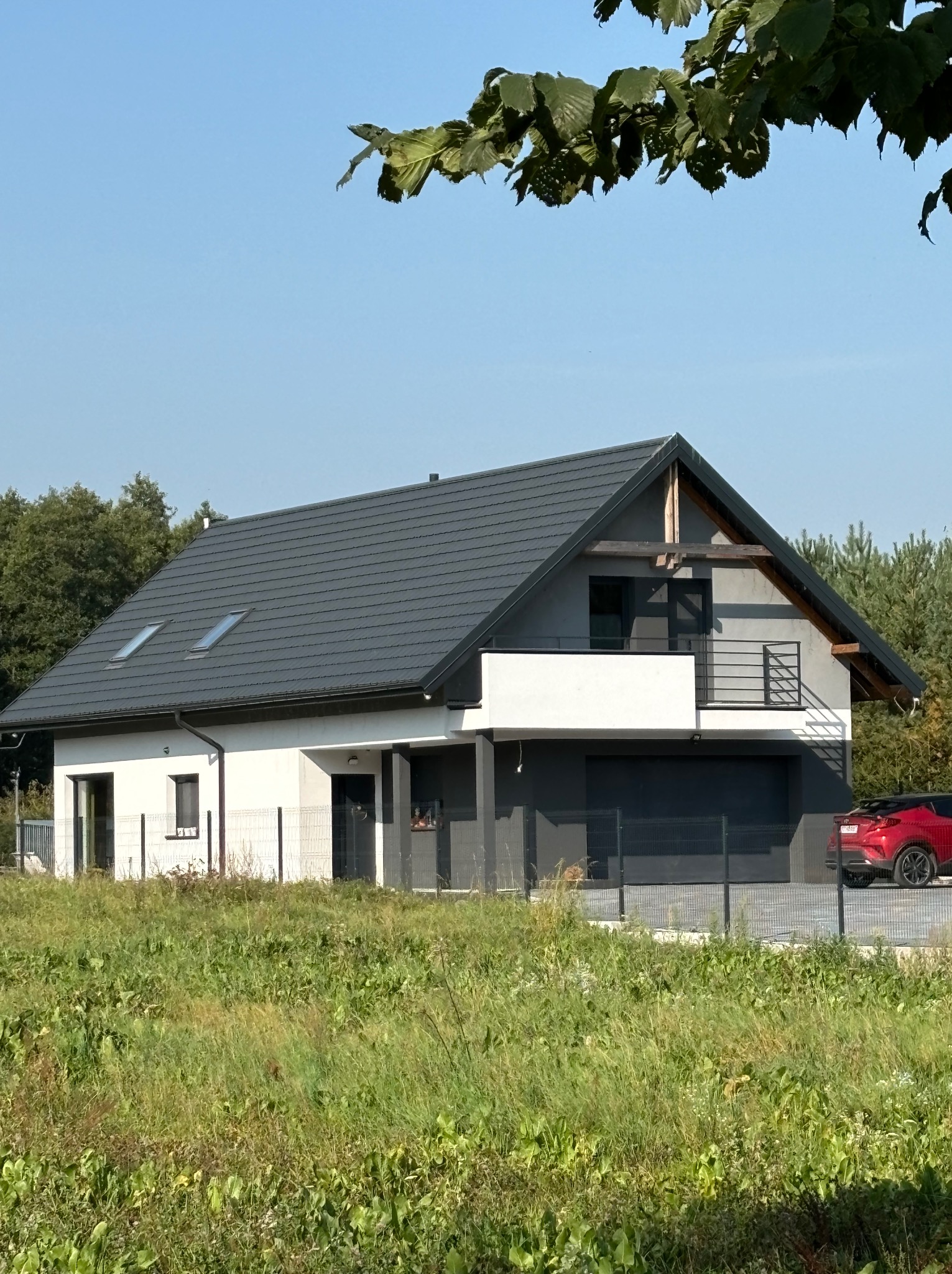 Nowoczesny dom jednorodzinny z szarą elewacją i dachem, widoczny zza wysokiej trawy, z czerwonym samochodem w tle.