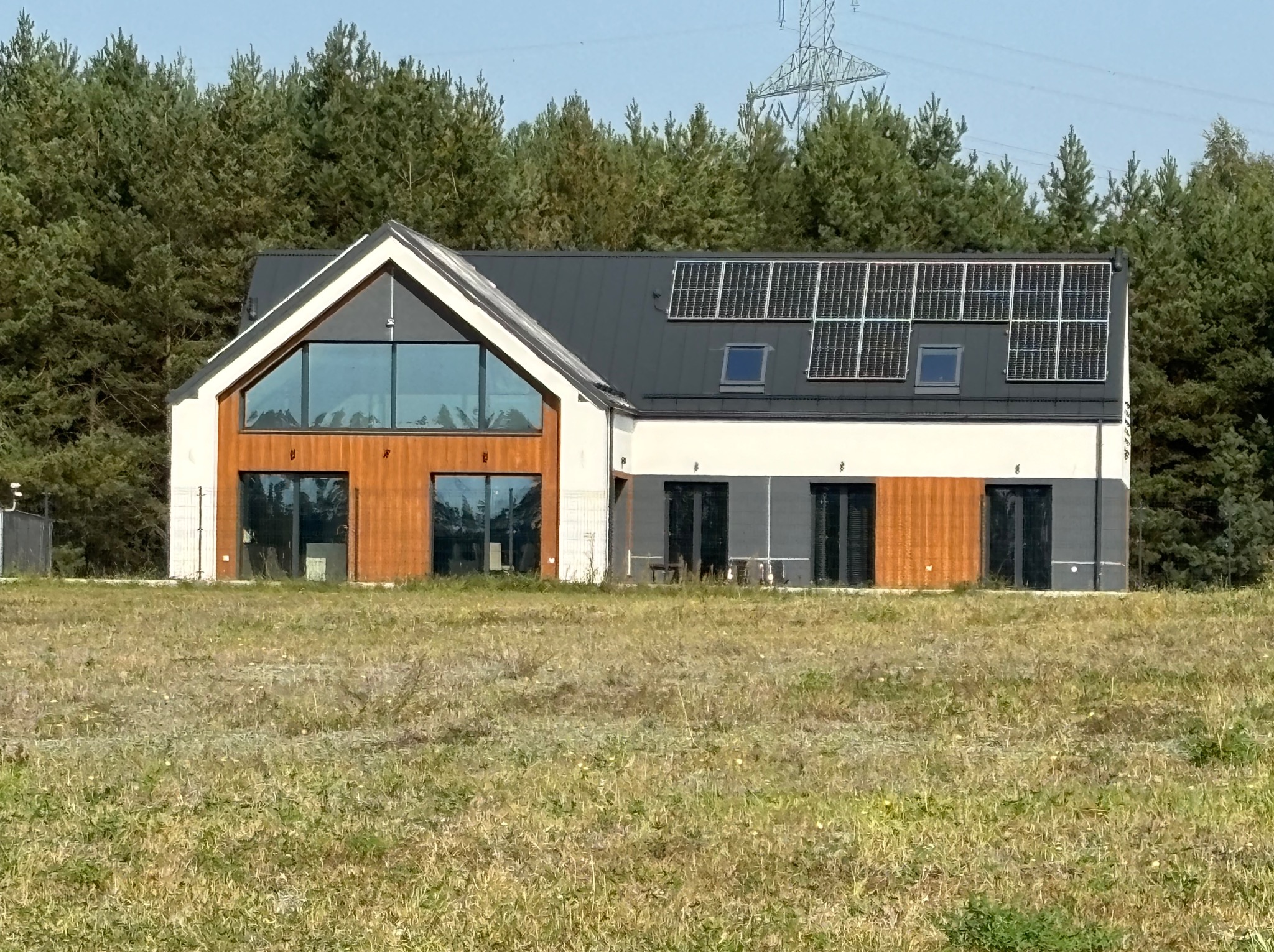 Nowoczesny dom jednorodzinny z panelami słonecznymi na dachu, wykończony drewnem i szarą elewacją, otoczony zielonymi drzewami i łąką.