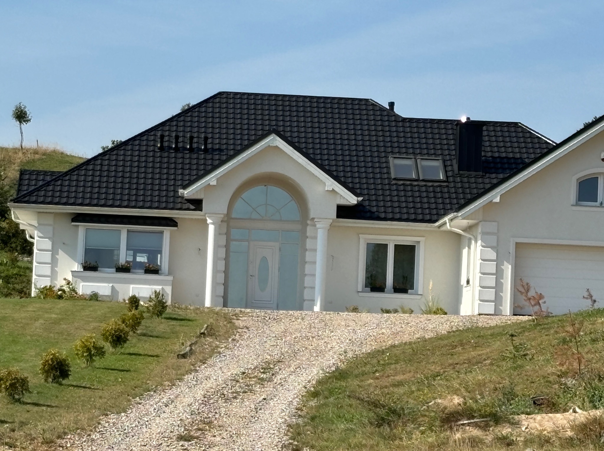 Elegancki dom jednorodzinny z białą elewacją, czarnym dachem z dachówki i podjazdem z jasnego żwiru, otoczony zielonym trawnikiem i niskimi krzewami.