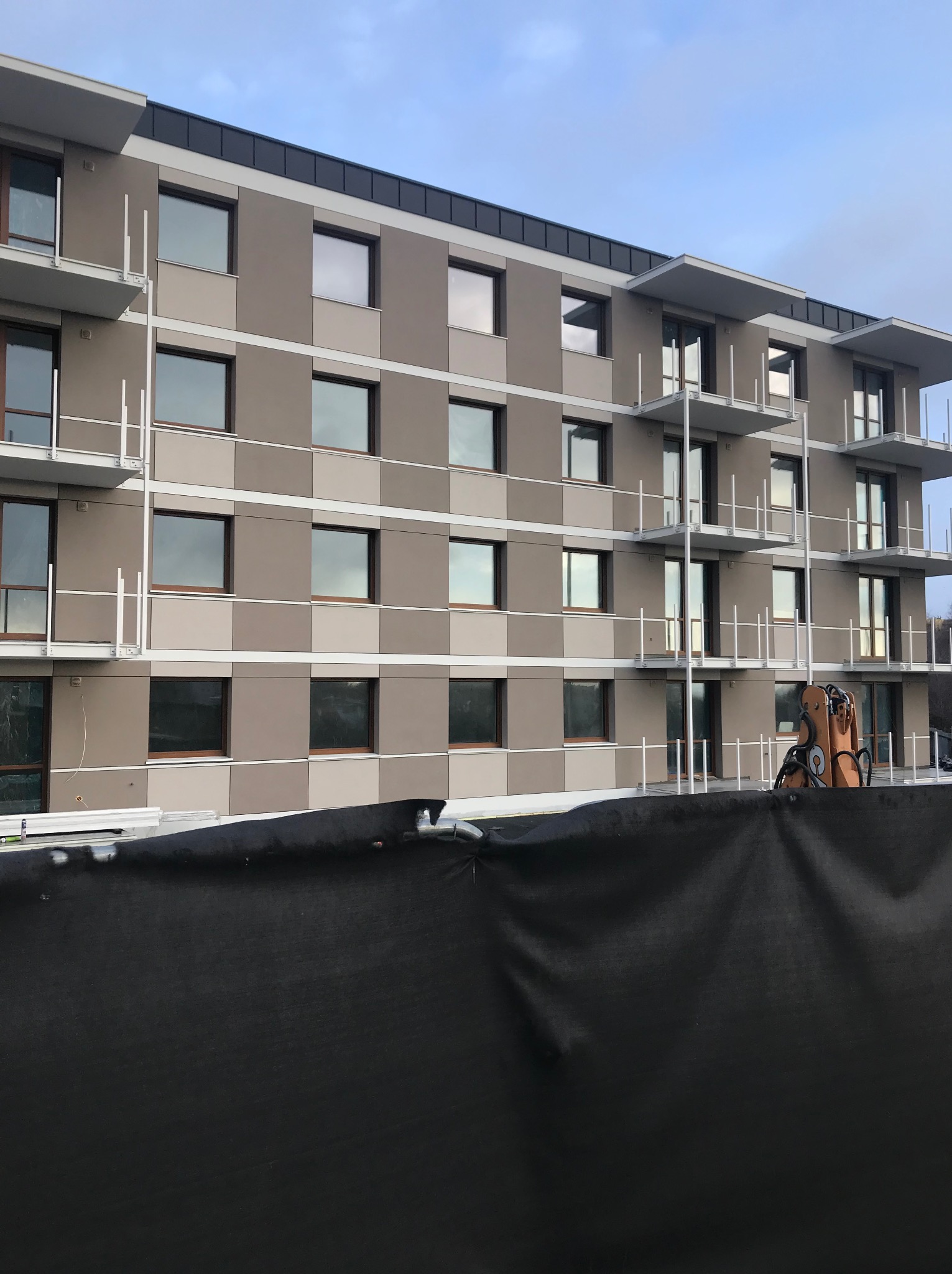 Nowoczesny budynek wielorodzinny z elewacją w odcieniach szarości, widoczny fragment czarnej siatki zabezpieczającej i sprzęt budowlany.