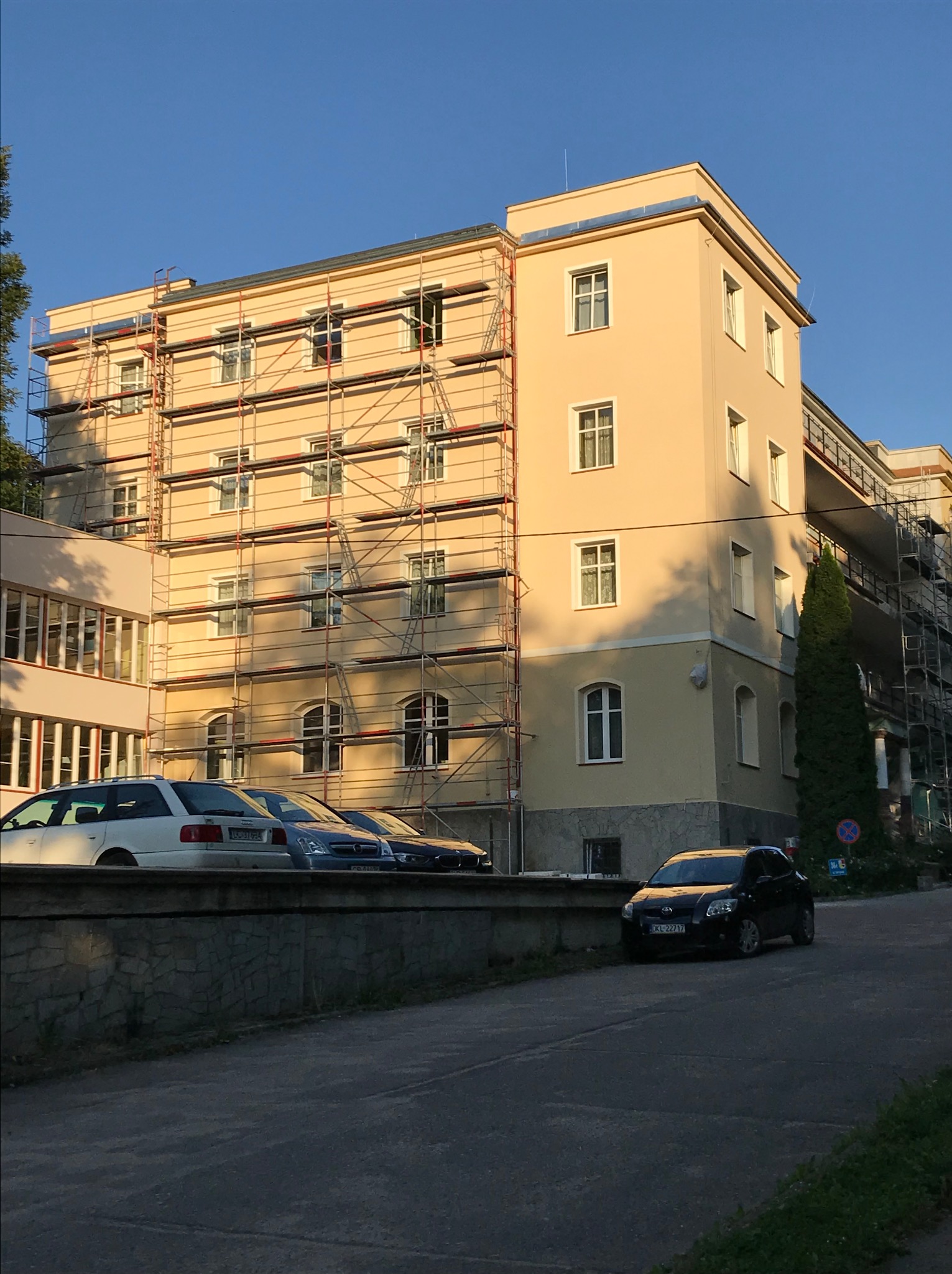 Budynek wielorodzinny w trakcie prac elewacyjnych, widoczne rusztowania, fragmenty ocieplenia i nowej elewacji, zaparkowane samochody w pobliżu.