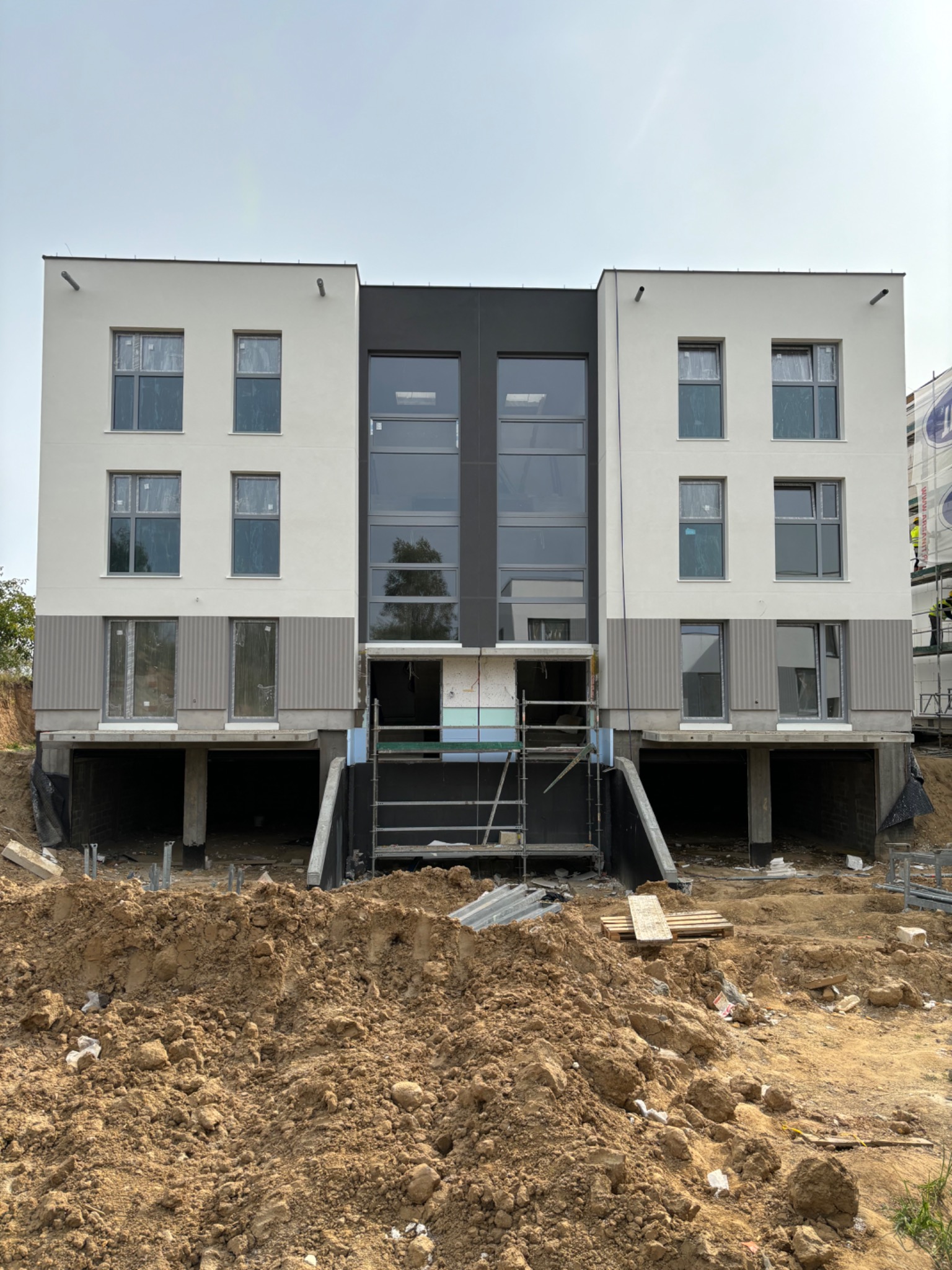 Dwukondygnacyjny budynek z szarą elewacją w trakcie prac wykończeniowych, widoczne rusztowania i ziemia przed budynkiem.