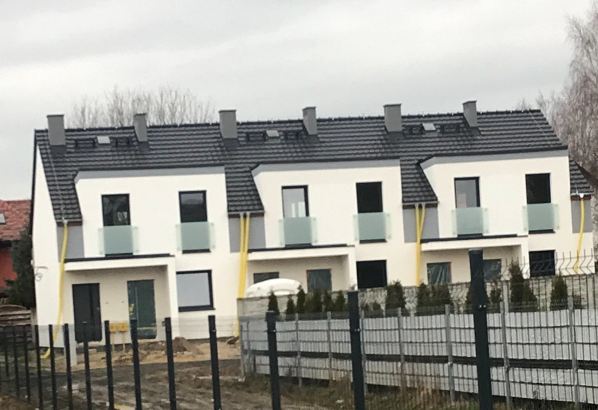 Szereg nowo wybudowanych, ocieplonych domów w zabudowie szeregowej z szarą dachówką i elewacją w kolorze złamanej bieli, widoczne żółte rury przy elewacji i ogrodzenie.