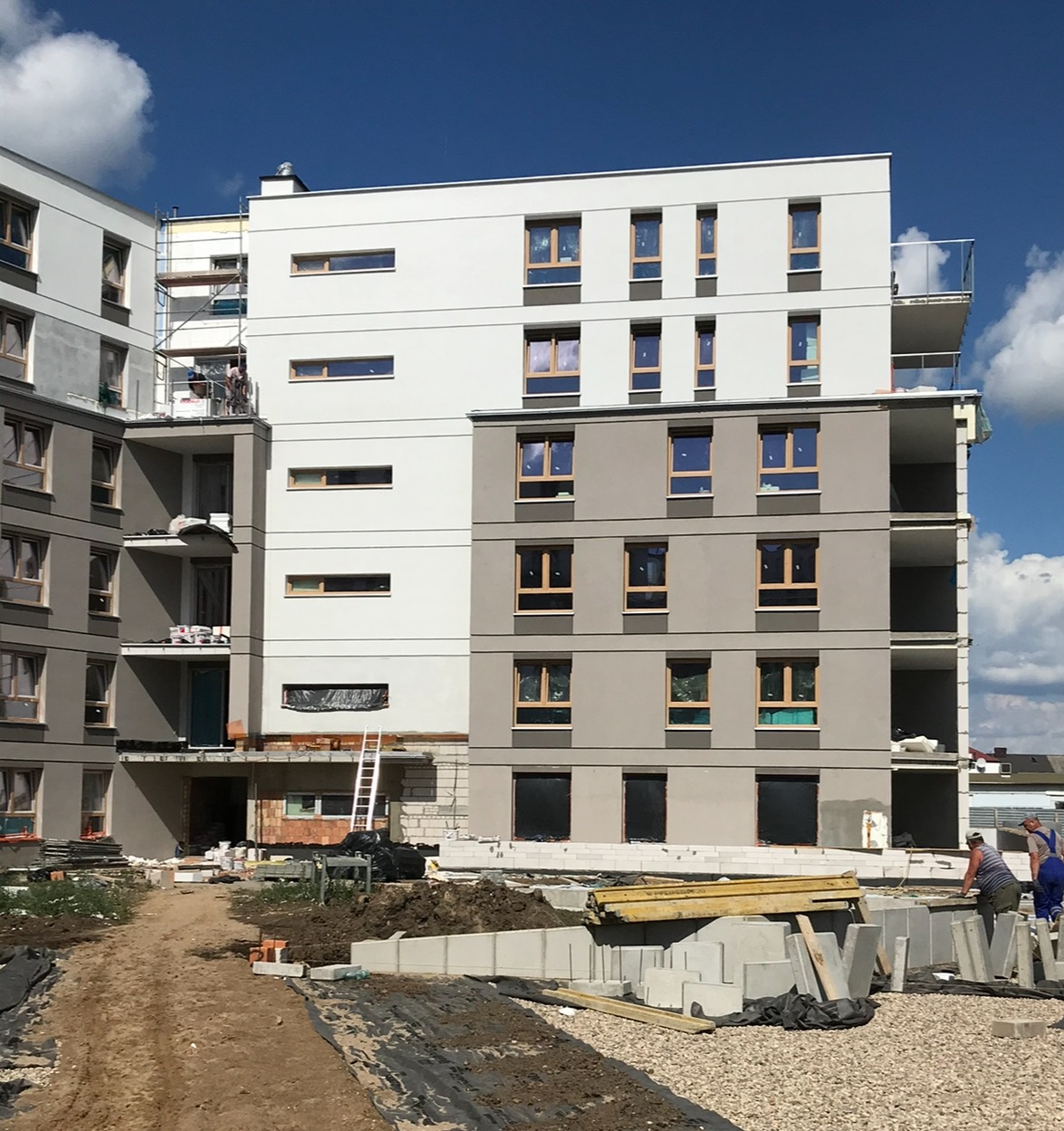 Wielopiętrowy budynek mieszkalny w trakcie wykańczania elewacji, widoczne rusztowania, materiały budowlane i pracownicy w oddali, częściowo pomalowane ściany w odcieniach bieli i szarości, błękitne...