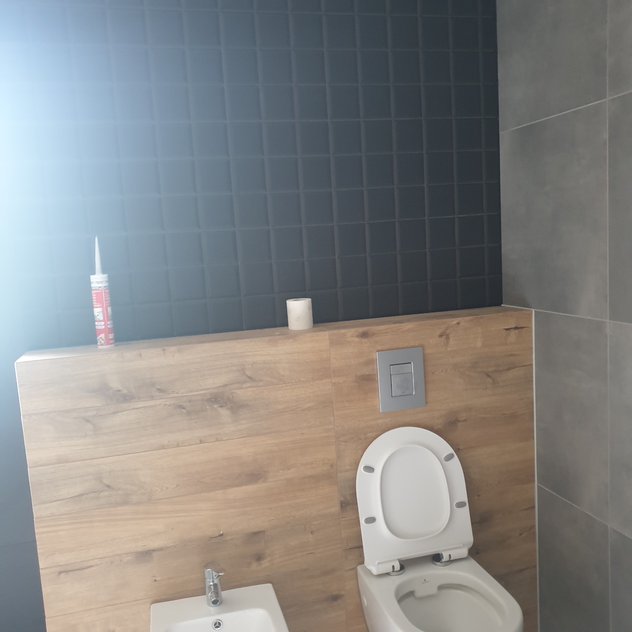 Nowoczesna łazienka z czarnymi płytkami mozaikowymi na ścianie, drewnopodobną zabudową stelaża, wiszącą miską WC i bidetem. Tubka silikonu i rolka papieru toaletowego leżą na zabudowie.