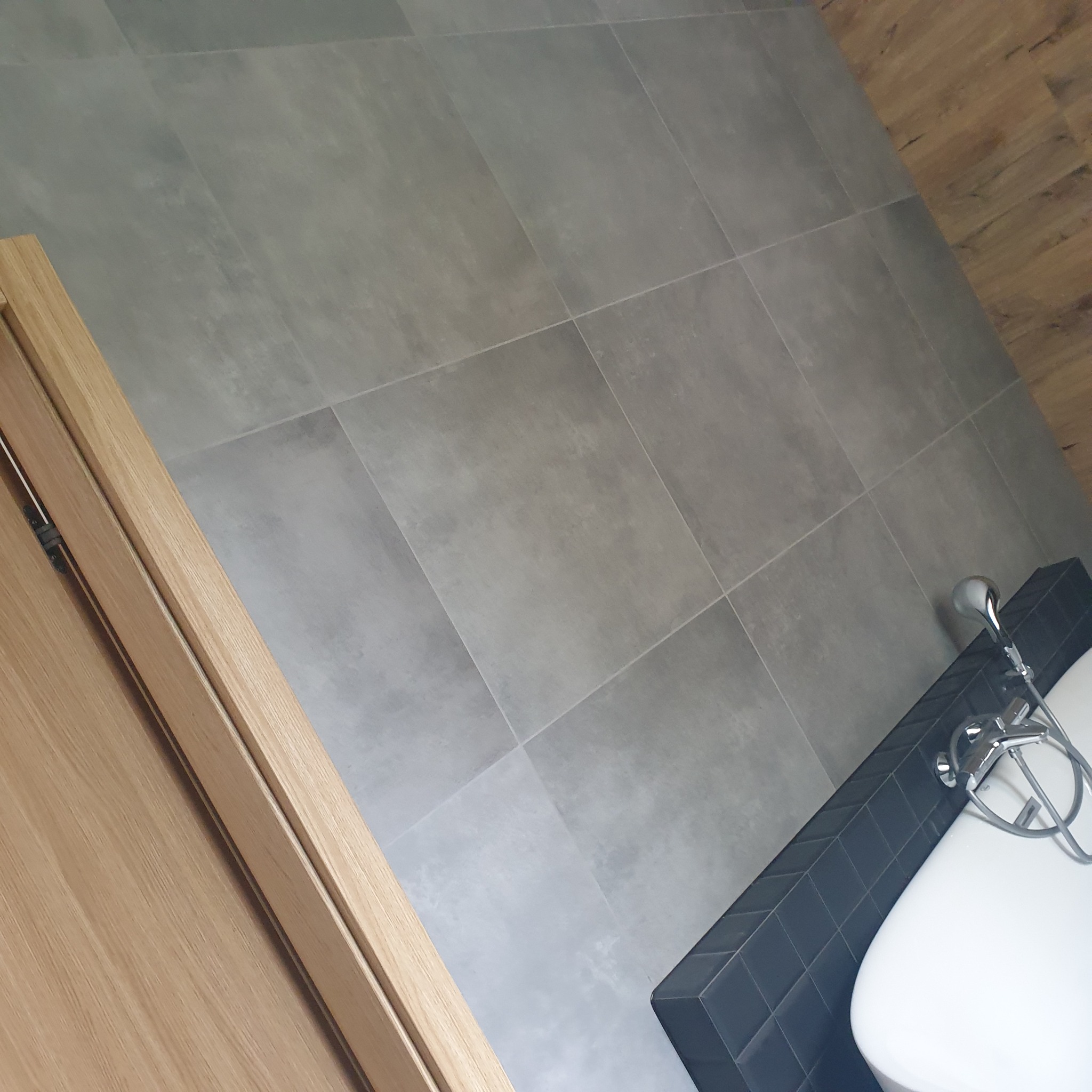 Widok z góry na fragment łazienki z szarą, kwadratową podłogą, drewnianą ościeżnicą drzwi po lewej stronie i fragmentem białej wanny z czarną obudową oraz chromowaną baterią w prawym dolnym rogu.