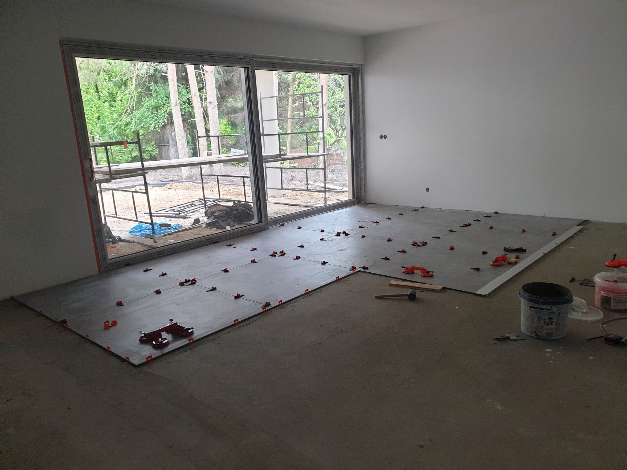 Przygotowanie do układania płytek podłogowych w pomieszczeniu z dużym oknem, widoczne poziomice i dystanse do płytek oraz wiadro z klejem.